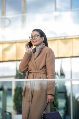 Bej ceketli gülümseyen Asyalı iş kadını dışarıda, modern iş merkezinin dışında dururken cep telefonuyla konuşuyor.