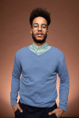 Zeki iş adamı, kahverengi arka planda elleri pantolonun cebinde kameranın önünde dikilirken sana bakıyor.