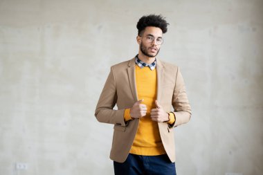 Genç, zarif, sıradan giyimli bir iş adamı kameranın önünde dikilip ceketine dokunurken sana bakıyor.