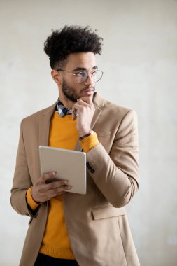 Genç ve düşünceli bir iş adamı, gri arka planda kameranın önünde dijital tablet kullanıyor.