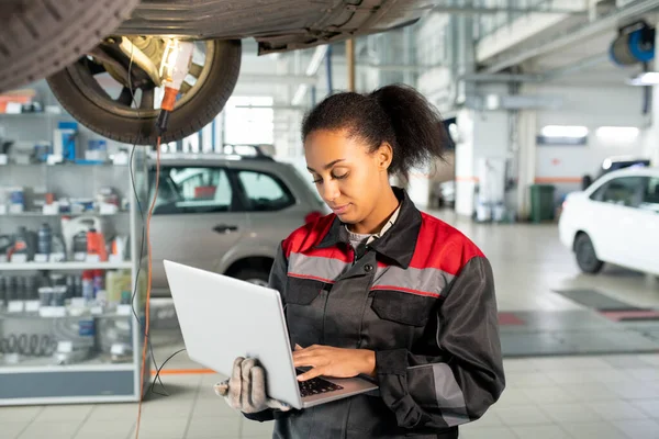 Female worker of car repair service in uniform using laptop in workshop to look for data in online sources, take new orders or consult clients