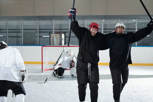 Two excited hockey players in sports uniform, gloves and protective helmets standing in front of camera after play and expressing joy