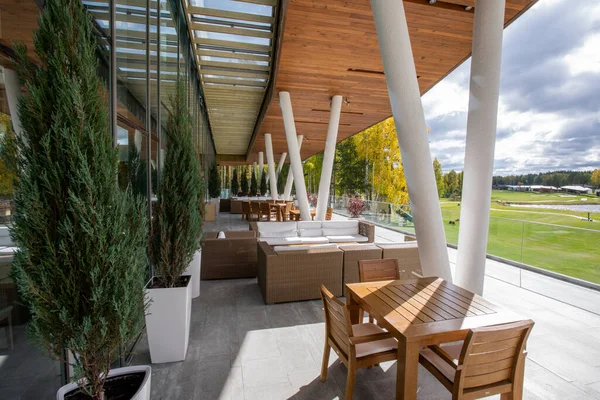 Wooden table and chairs, couches and green decorative trees growing in pots standing outdoors along large windows of modern business center