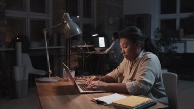 Ağır çekimde, yorgun Afrikalı Amerikalı olgun bir kadının karanlık ofis odasında oturmuş laptopta son teslim tarihlerini vurgularken görüntüsü.