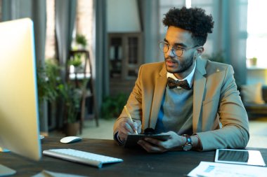 Genç işadamı masada oturup bilgisayar ekranına bakıyor. İnternetteki iş görüşmesi sırasında notlar alıyor.