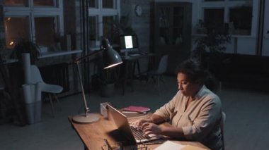 Yüksek açılı, siyah, boş bir çatı katında, dizüstü bilgisayarın önünde oturan, geceleri yalnız çalışan bir afro kadının fotoğrafı.