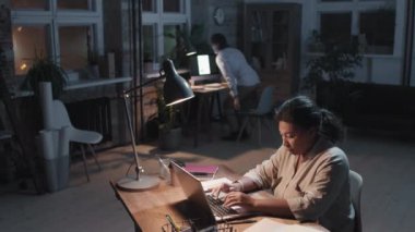 Yüksek açılı, ağır çekimde, çok ırklı iki meslektaşın geç saatlere kadar görevde kalması, erkeğin işini bitirip gitmesi ve kadının hala işine devam etmesi.