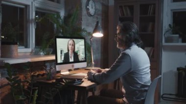 Yetişkin bir adamın karanlık bir ofiste bilgisayar monitörünün önünde iş görüşmeleri yaparken orta açıdan görüntüsü.