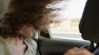 Uzun kıvırcık saçlı mutlu beyaz kadın arabayla seyahat ederken şarkı söyleyip dans ediyor.