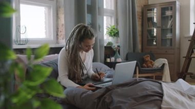 Rasta giymiş, evinde oturmuş, laptopuna soğutulmuş atmosferde bağlı tablet çizen genç ve güzel bir kadının yavaş çekim görüntüsü.