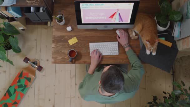 オンラインショッピングをしながらお茶を飲みながら 赤い猫とテーブルに座っている若い男のトップビューの映像 — ストック動画