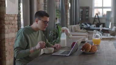 Masada oturup laptopla kahvaltı yapan serbest çalışan bir erkeğin orta ölçekli yan görüntüsü.