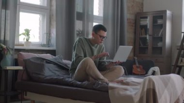 Ağır çekimde, beyaz dövmeli, yatakta oturmuş, parlak bir dairede dizüstü bilgisayarla çalışırken çekilmiş.