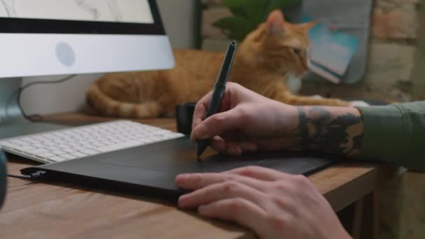 ゆっくりとクローズアップの認識できない入れ墨男性グラフィックデザイナードローイングオンタブレットでペンと一方 シックな赤猫冷たいですデスクで背景 — ストック動画