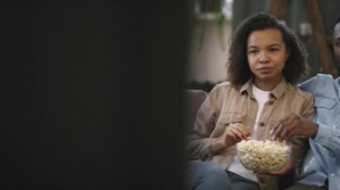 Yavaş çekimde, parlak ve rahat oturma odasında TV karşısında oturan, patlamış mısır yiyen ve film izlerken film tartışan genç afro çiftin belden yukarı çekimleri.