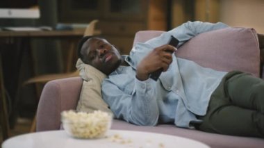 Orta boy, tembel, Afrikalı Amerikalı bir adamın gece televizyon karşısında kanalı değiştirip uzaktan kumandayla uykuya daldığı bir fotoğraf.