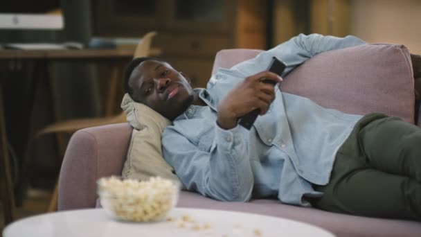 夜にはテレビの前でソファに横たわっ若い怠惰なアフリカ系アメリカ人の男の中のショット その後 手でリモートコントロールで眠りに落ちるチャンネルを切り替える — ストック動画