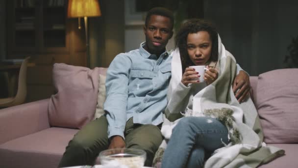 年轻的爱情非洲裔情侣晚上一起坐在沙发上看电影的慢镜头镜头 女人生病了 喝着覆盖着格子花的热茶 而男人拥抱着她 — 图库视频影像