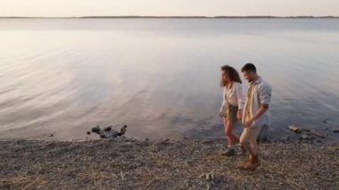 Yüksek açılı, birbirine aşık genç adam ve kadının akşam vakti güzel göl kıyısında yürürken görüntüsü.