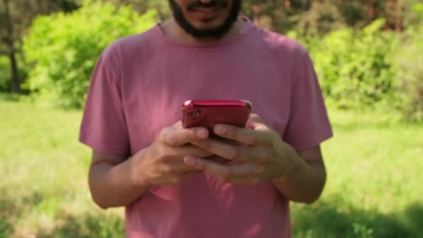 手の中にピンクのTシャツを保持スマートフォンを身に着けている認識できない髭の男の中セクションのショットサーフィンインターネット — ストック動画