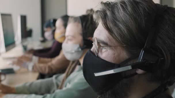 戴着面具和耳机在呼叫中心工作并回答询问的年轻人的近照 — 图库视频影像
