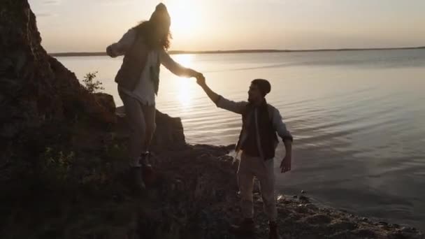 恋人を助ける彼女から降りて岩に沿って行く湖の海岸一緒に手を保持し バックライトショット — ストック動画