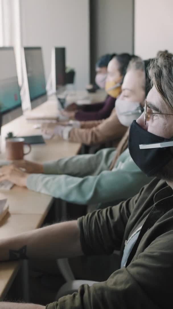 パノラマウィンドウで机の上に座っている顔のマスクやヘッドセットの人々の垂直Panショットやコンピュータ上のオーディオコールでお問い合わせに答える — ストック動画