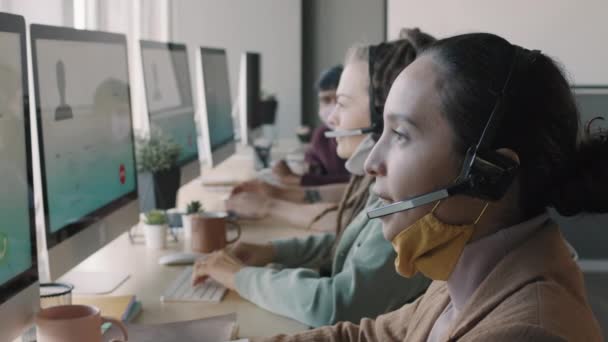 年轻电话中心工作人员头戴耳机和面罩 坐在电脑显示器前帮助客户或进行销售时 与Pan的慢速侧视 — 图库视频影像