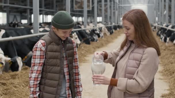 中镜头的快乐女子站在奶牛饲养场的奶牛场里 把鲜牛奶倒入杯子里 快乐地看着男孩笑着 — 图库视频影像