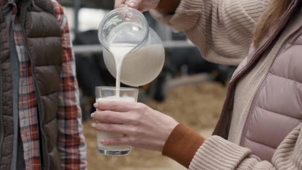 关闭中区Pan无法辨认的妇女倒入新鲜牛奶的杯子 并把它交给青少年男孩 饲养场的奶牛在后方吃干草 — 图库视频影像