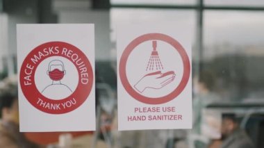 Yüz Maskeleri Gereklidir yazan tabelaların PAN görüntüsü ve ofis binasının içindeki cam duvarda lütfen el dezenfektanı kullanın.