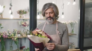 Küçük bir çiçekçide çiçekçi olarak çalışan, elinde güzel bir buket çiçek tutarken kameraya gülümseyen, beyaz tenli yetişkin bir adamın yavaş çekim belindeki portresi.