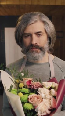 Dikey yavaş çekim orta boy profesyonel yetişkin çiçekçinin güzel bir buket çiçek tutup kameraya baktığı portresi.