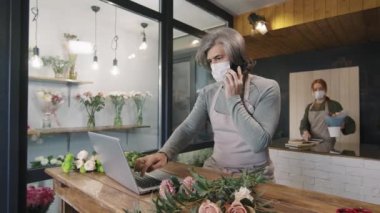 Yetişkin erkek çiçekçinin koruyucu maske taktığı, telefonda sohbet ettiği ve dizüstü bilgisayarla küçük bir çiçekçide tezgahta durduğu orta boy bir fotoğraf.