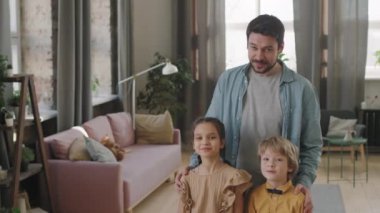 Kafkasyalı mutlu bir adamın ve iki çocuğunun, modern, rahat oturma odasında kameraya gülümseyen iki çocuğunun yavaş çekimde orta ölçekli portresi. Aile ve ebeveynlik kavramı