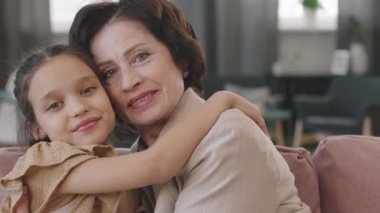 Yavaş çekimde orta boy PAN portresi. Güzel görünümlü yaşlı bir kadın. Küçük güzel bir kıza sarılıyor, kanepeye oturuyor ve kameraya gülümsüyor.