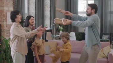 Kadınlar Günü 'nde çok nesilli kadınları tebrik eden hediyeleri olan neşeli bir adamla küçük bir çocuğun orta boy PAN resmi. Çocuklar hediye veriyor, herkes sarılmış ve gülüyor oturma odasında.