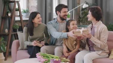 Uluslararası Kadın Günü 'nü kutlayan mutlu ve büyük bir ailenin yavaş çekimleri. Erkek, güzel yetişkin kadına hediyeler ve çiçekler veriyor. Oturma odasında kanepede yan yana oturuyor.