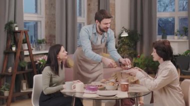 Bir beyefendinin yemek masasında kadınlarla ilgilenirken, tabağa bir parça kek koyarken ve gülümserken orta ölçekli bir resmi. Herkes Uluslararası Kadınlar Günü 'nü kutluyor.