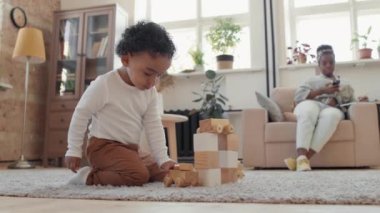 Oturma odasında halıya diz çöküp ahşap yapı taşları ve tren setiyle oynayan sevimli zenci çocuğun yavaş çekimleri. Annesi kanepede oturup cep telefonu kullanıyor.