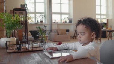 Kıvırcık saçlı, sevimli bir çocuğun orta boy fotoğrafı. Masasında oturmuş, rahat bir dairede tablet üzerinde çizgi film seyrediyor. Annem çerçeveye giriyor ve arka planda oyuncaklarını topluyor.