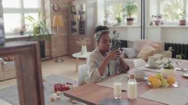 Genç siyahi bir kadının, havadar bir dairede yemek masasında oturup sabahları sıcak çay içerken çekilmiş el kamerasıyla çekilmiş bir fotoğrafı.