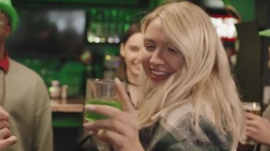 St. Patrick Günü 'nü kutlayan çok ırklı arkadaşlarla birlikte İrlanda barında kameraya dans ederek dans eden genç ve güzel bir sarışınla orta boy yakın çekim.