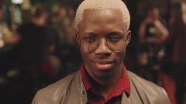 Gözlüklü, boyalı sarı saçlı ve akşamları barda eğlenirken gülümseyen kameraya bakan Afro-Amerikan 'ın yavaş yavaş çekilmiş yakın plan portresi.