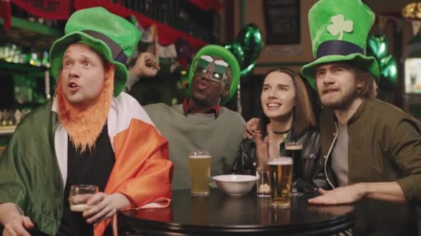 在圣帕克斯节当天 一群兴奋的多民族朋友在当地爱尔兰酒吧的电视上观看体育比赛 他们的慢镜头闪烁着兴奋的光芒 一个接一个地欢呼着 — 图库视频影像