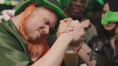 Yeşil İrlanda şapkalı neşeli adamın yavaş çekimleri yerel bir barda otururken bilek güreşi yapması ve arkadaşlarının izlemesi, herkesin eğlenmesi.