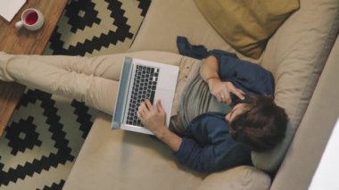 Kanepede bacaklarını sehpaya dayamış, cep telefonuyla konuşurken dizüstü bilgisayara bakan genç bir adamı izliyoruz.