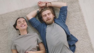Genç kadın ve erkeğin gözleri kapalı, gülümseyen ve halıya uzanan görüntüsü.