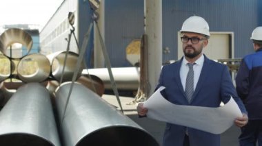 Çelik boruların yanında duran takım elbiseli ve kasklı ciddi erkek imalat mühendisinin yavaş PAN 'ı planlara bakıyor. Arka planda yürüyen üniformalı işçiler