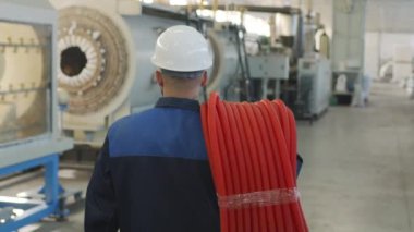  Kırmızı HDPE borularını taşıyan ve fabrika tesisinde üretim ekipmanlarıyla yürüyen üniformalı ve kasklı erkek işçilerin dikiz aynasından yavaş izleme.